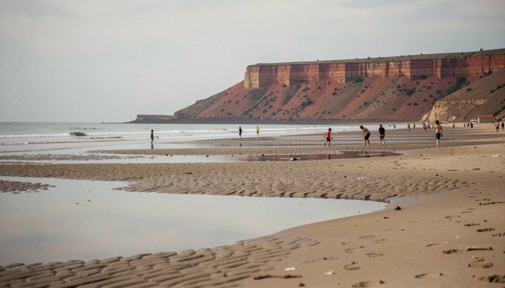A wide sandy beach at low tide with shallow pools, a distant red cliff and children playing near the water.